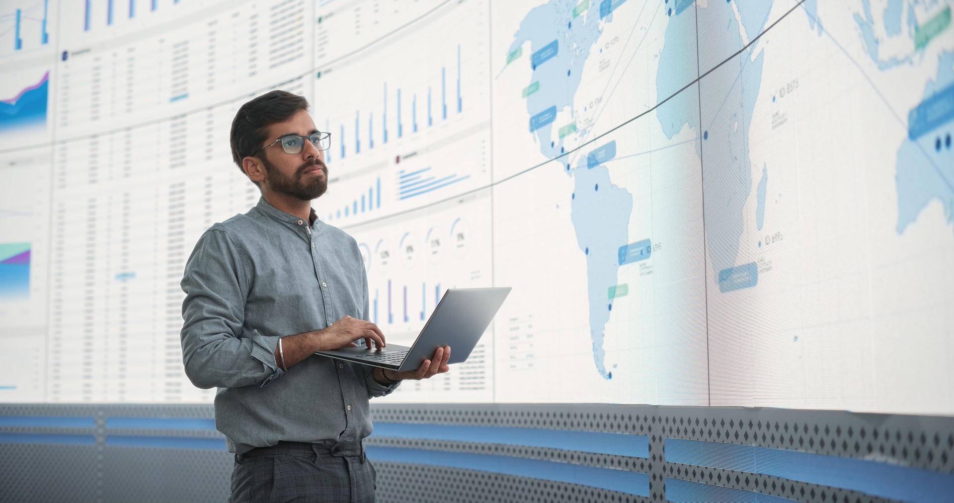 Experto en logística masculino indio que sostiene una computadora portátil y analiza el mapa del mundo en una gran pantalla digital en la oficina de monitoreo. Hombre exitoso desarrollando nuevas rutas eficientes para la distribución global de productos